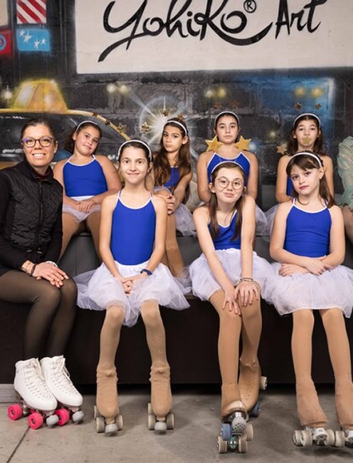 Patineuses artistiques en justaucorps bleu avec Yohiko Art Groupe de patineuses artistiques en justaucorps bleu et tutu blanc, posant avec leur entraîneur devant une fresque murale "Yohiko Art".