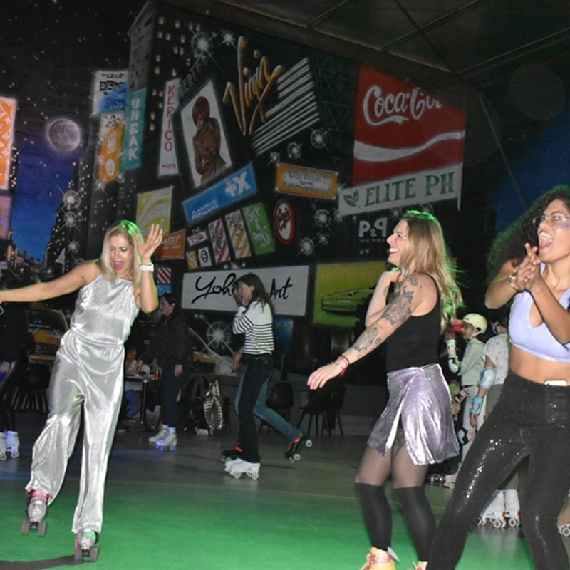 Soirée Roller Disco : Femmes joyeuses et fresque New York Quatre femmes souriantes et excitées patinent en rollers, l'une en combinaison argentée, sur une piste verte devant une fresque murale New York avec logos "Virgin" et "Coca-Cola".