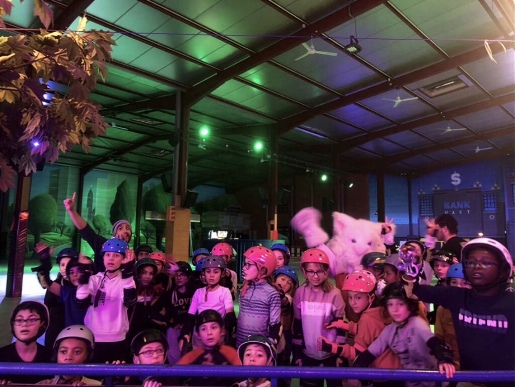 Enfants en casques et mascotte géante à la patinoire intérieure. Groupe d'enfants en casques avec une mascotte blanche géante. Un adulte pointe. Décor intérieur d'une patinoire avec l'enseigne "BANK WALT".