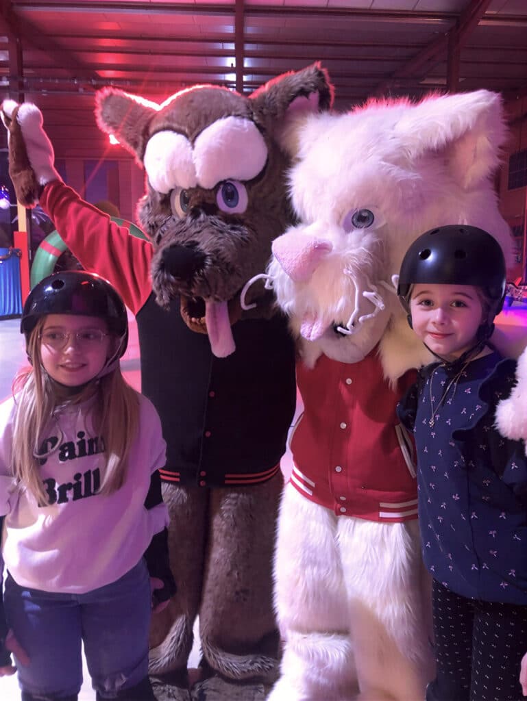 Rencontre de mascottes : loup et chat avec enfants en casque. Deux filles avec casques posent entre des mascottes géantes (loup brun, chat blanc). Sweat : "Pain Brill". Lumière rouge, joyeux moment.
